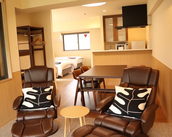 A modern hotel room with a dining area, two leather chairs with patterned cushions, a small round table, a bed with a white comforter, and a TV mounted above the bed. - Condominium Hotel Garland Court Usami (Ito)