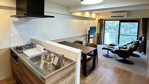 A modern kitchen with a built-in stove, a sink, and a black range hood. There is a dining area with a table and chairs, and a large window offering a view of the outdoors. - Condominium Hotel Garland Court Usami (Ito)