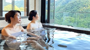 Two people in a hot tub with a view of a mountainous landscape through large windows. - Condominium Hotel Garland Court Usami (Ito)