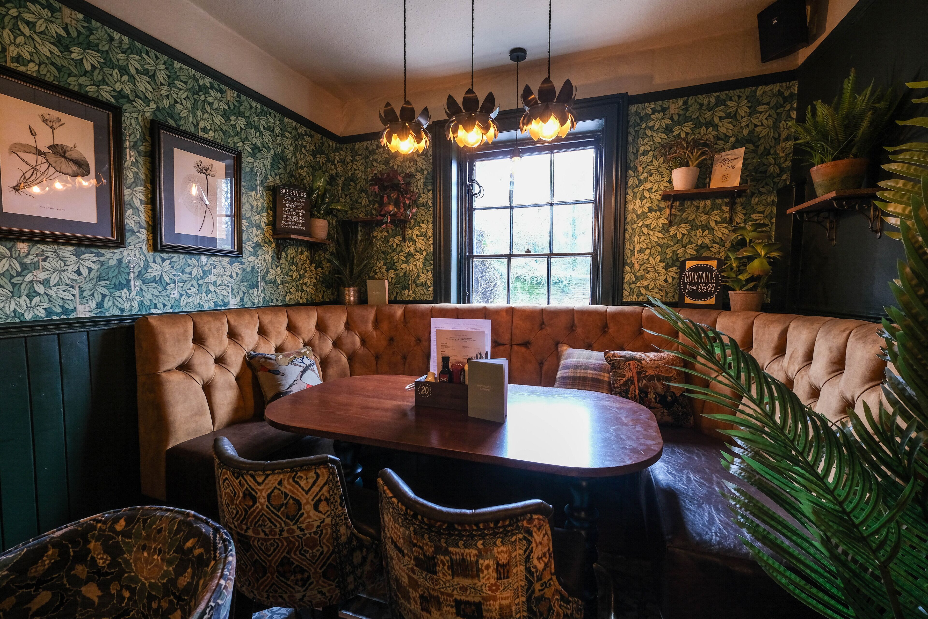 A cozy restaurant with a round wooden table, cushioned booth seating, patterned chairs, and a green leafy wallpaper.
