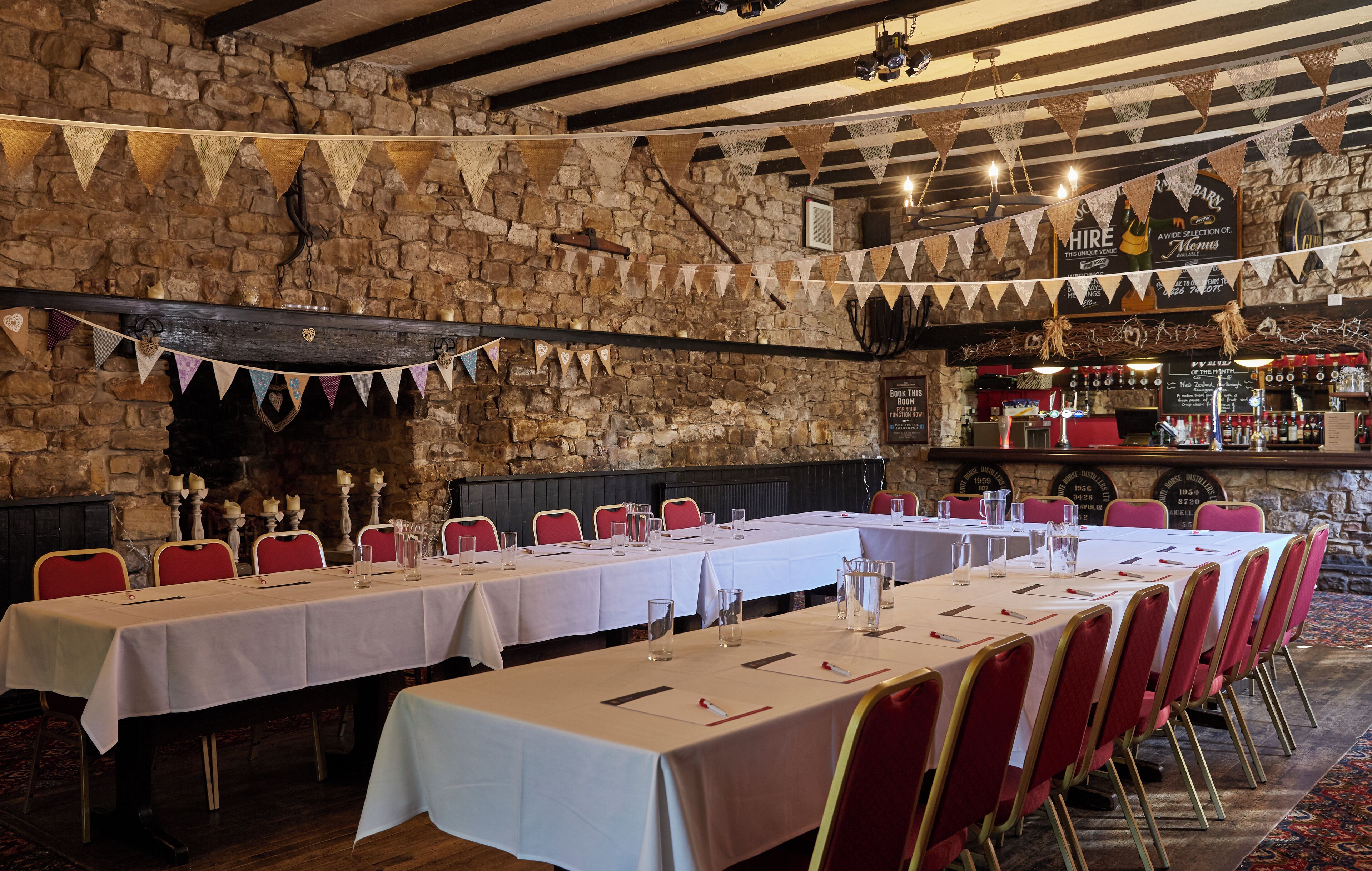 Un restaurant aux murs de pierre, aux chaises rouges et aux tables blanches. Des banderoles sont suspendues au plafond, et un bar est aménagé avec des bouteilles et des verres.