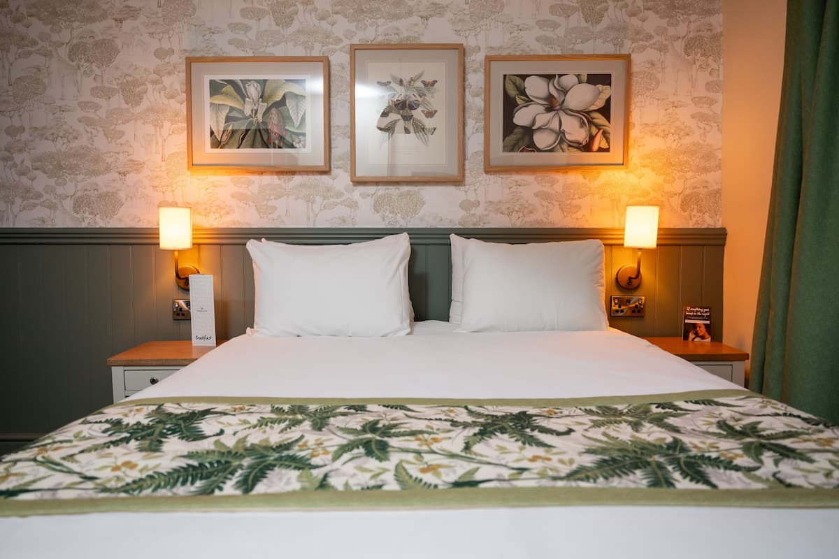 A neatly made bed with white linens, flanked by two bedside lamps, and framed botanical prints on the wall above.