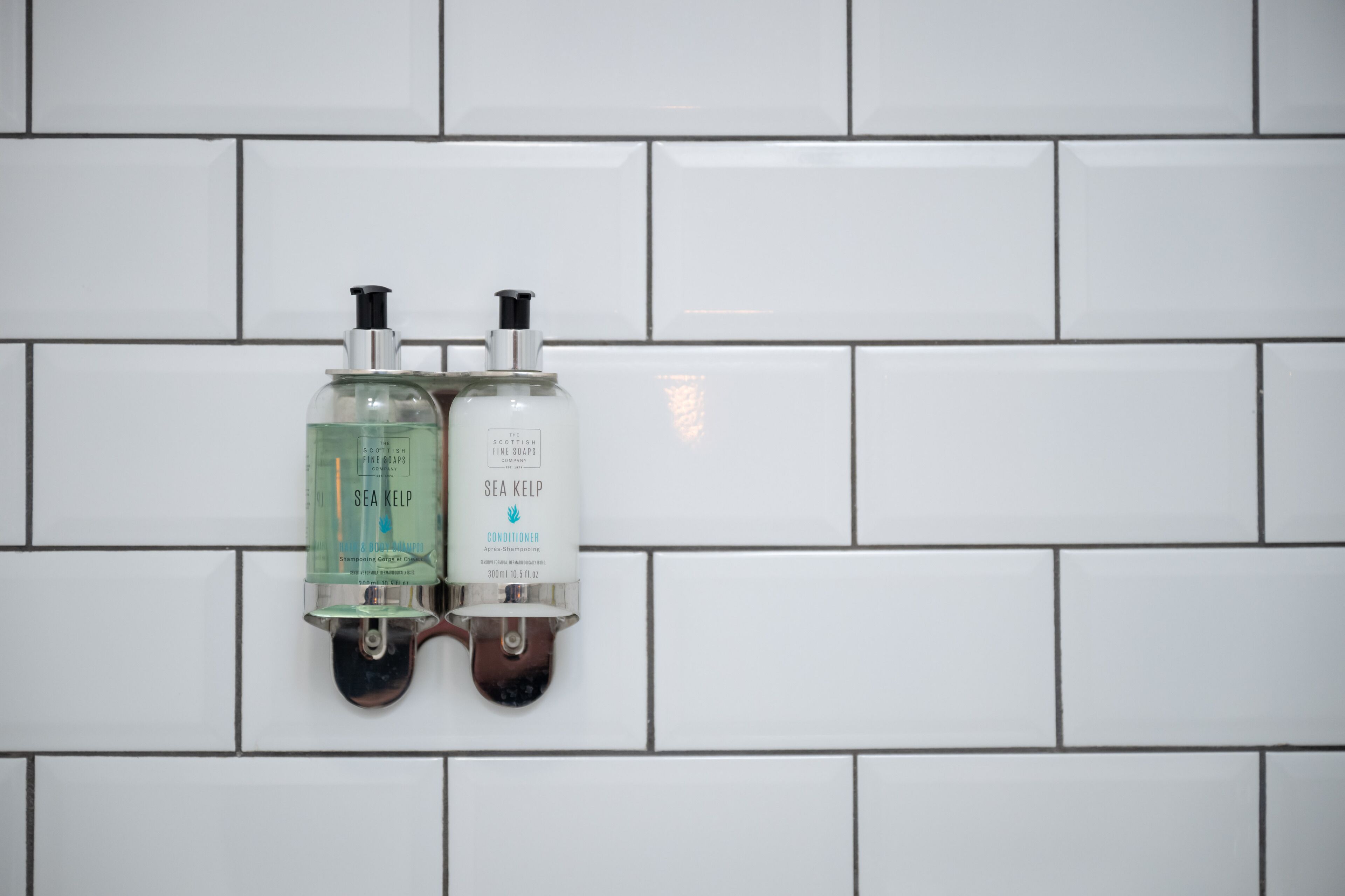 Dos dispensadores de jabón de manos fijos en una pared de azulejos blancos.