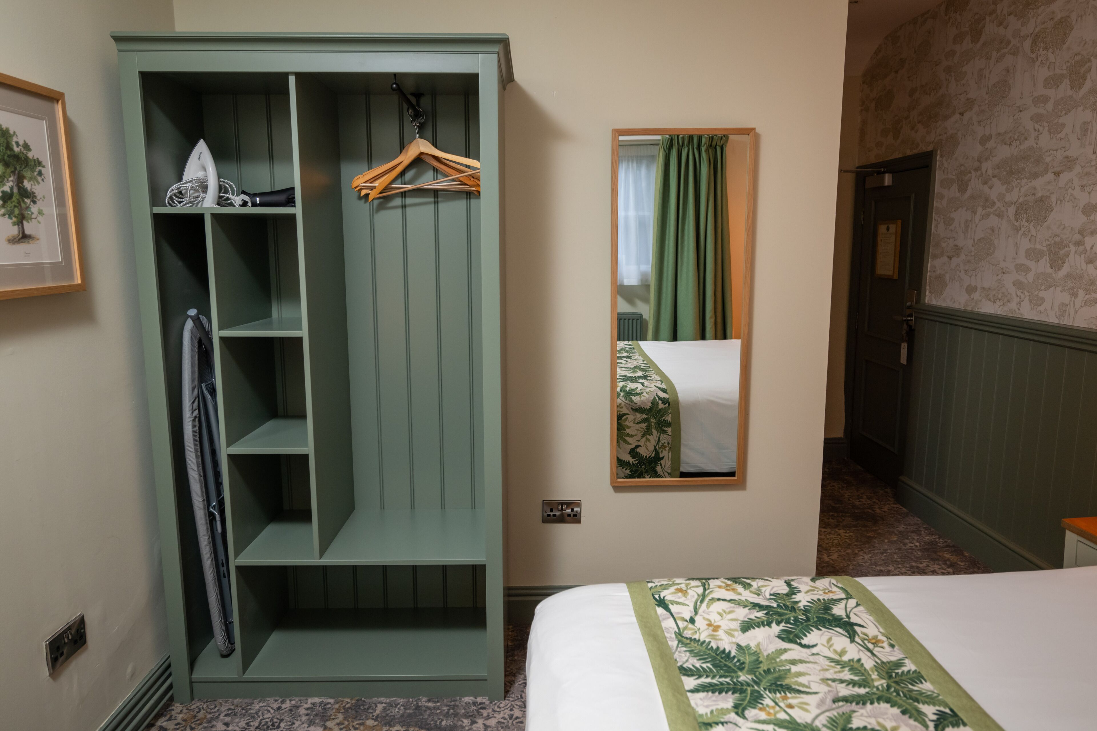 Ein Hotelzimmer mit einem grünen Kleiderschrank, einem Bett mit floralem Überwurf, einem Spiegel mit grünem Vorhang und einem gerahmten Bild an der Wand.