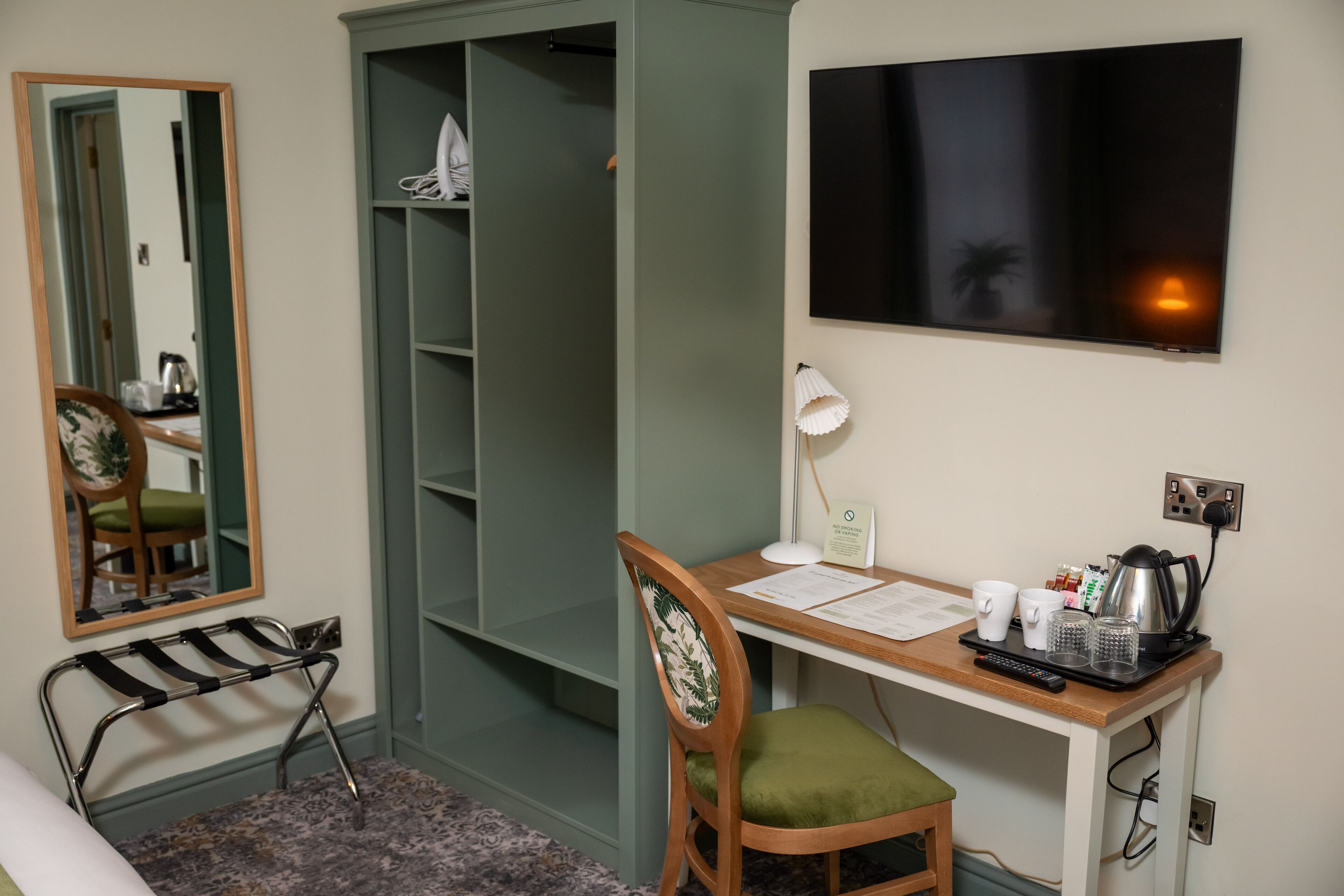 A room with a desk, chair, mirror, wardrobe, and a mounted TV.