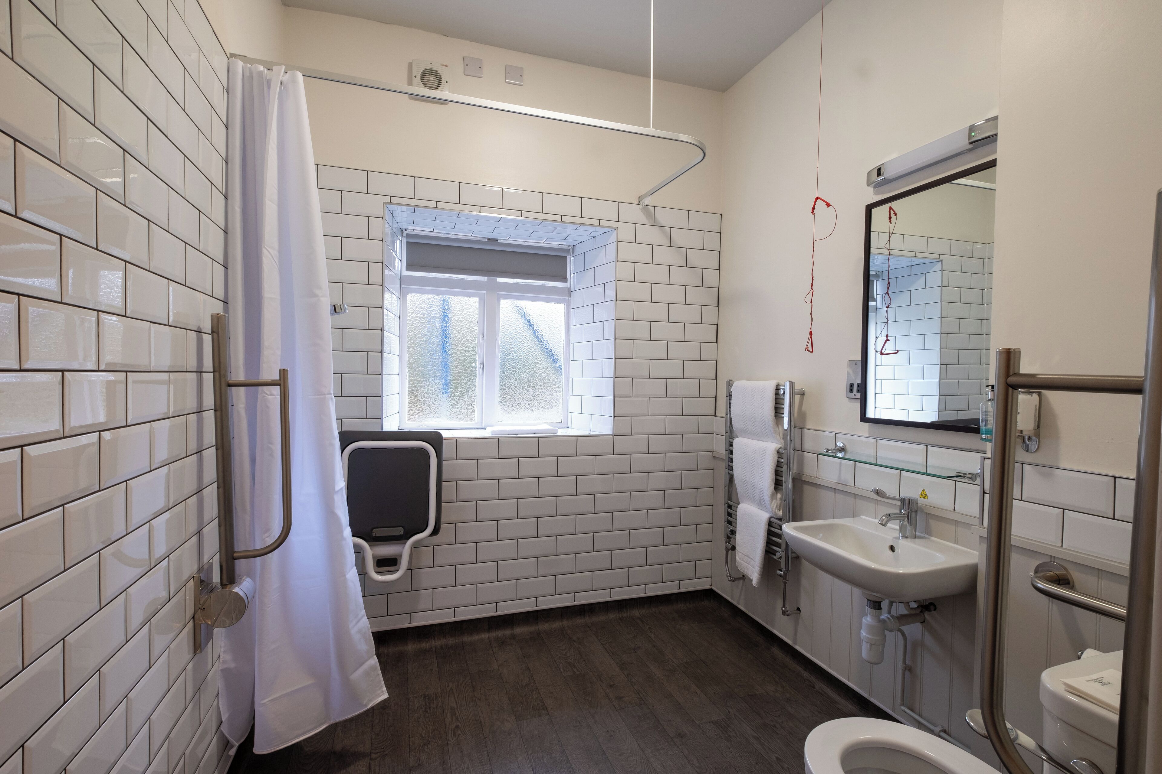 Un baño con paredes de azulejos blancos, una ventana, ducha, lavamanos e inodoro.