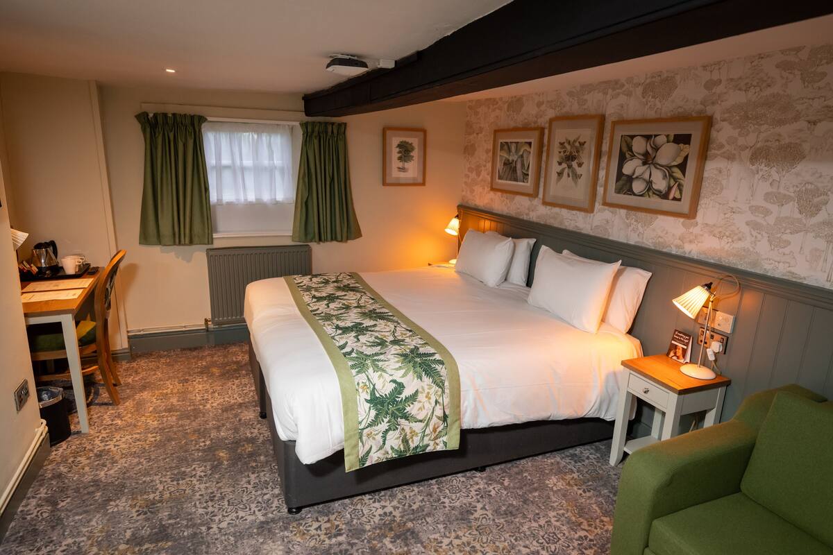 A hotel room with a large bed, a desk, a chair, and a nightstand with a lamp.