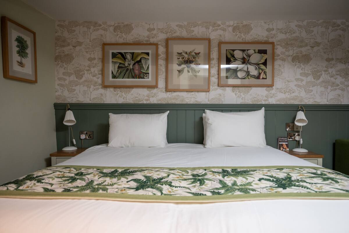 A neatly made bed with a floral bedspread, a green headboard, and framed botanical prints on the wall.