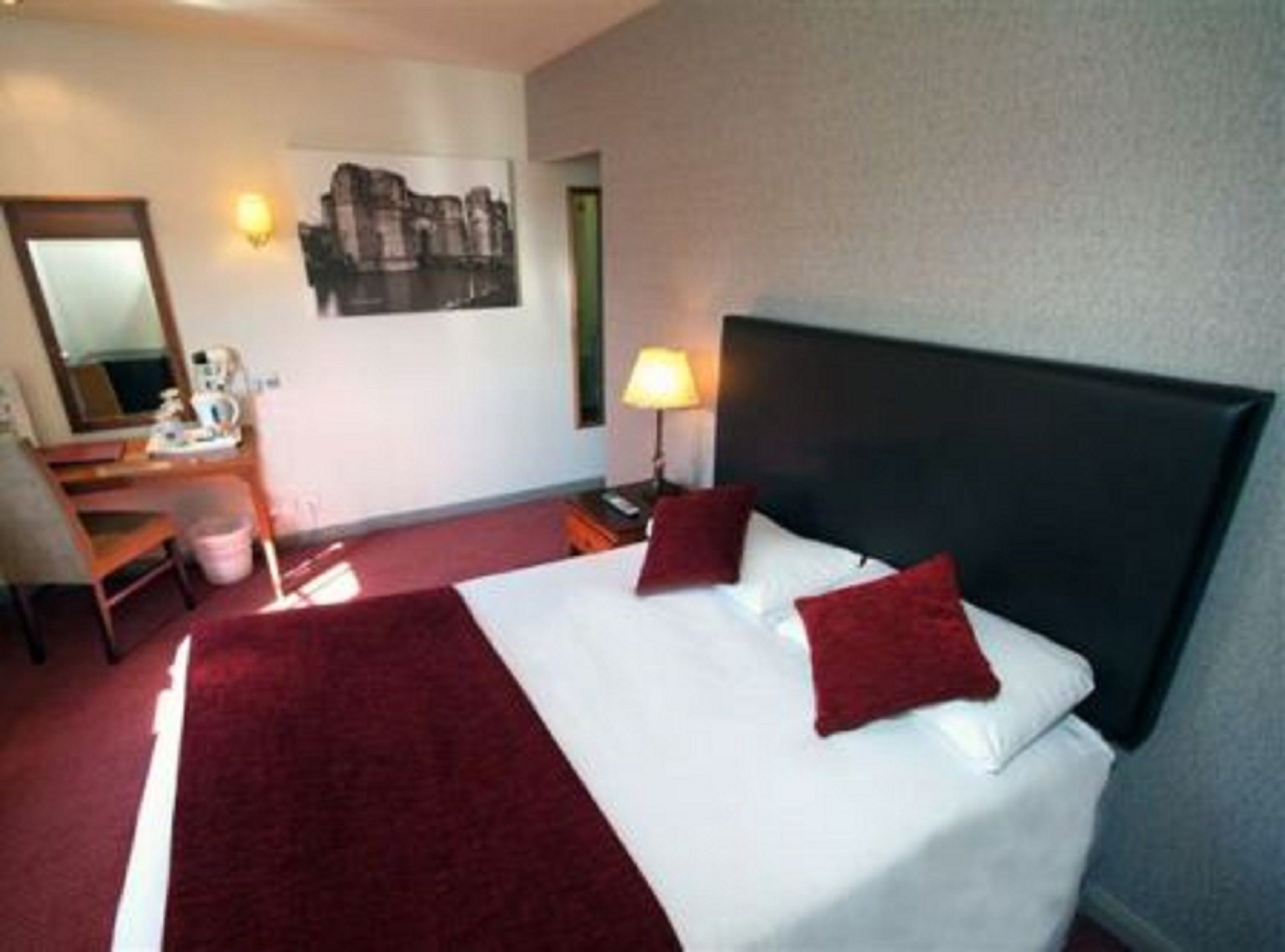 Una habitación de hotel con una cama grande, un escritorio con silla, una lámpara y un espejo montado en la pared.