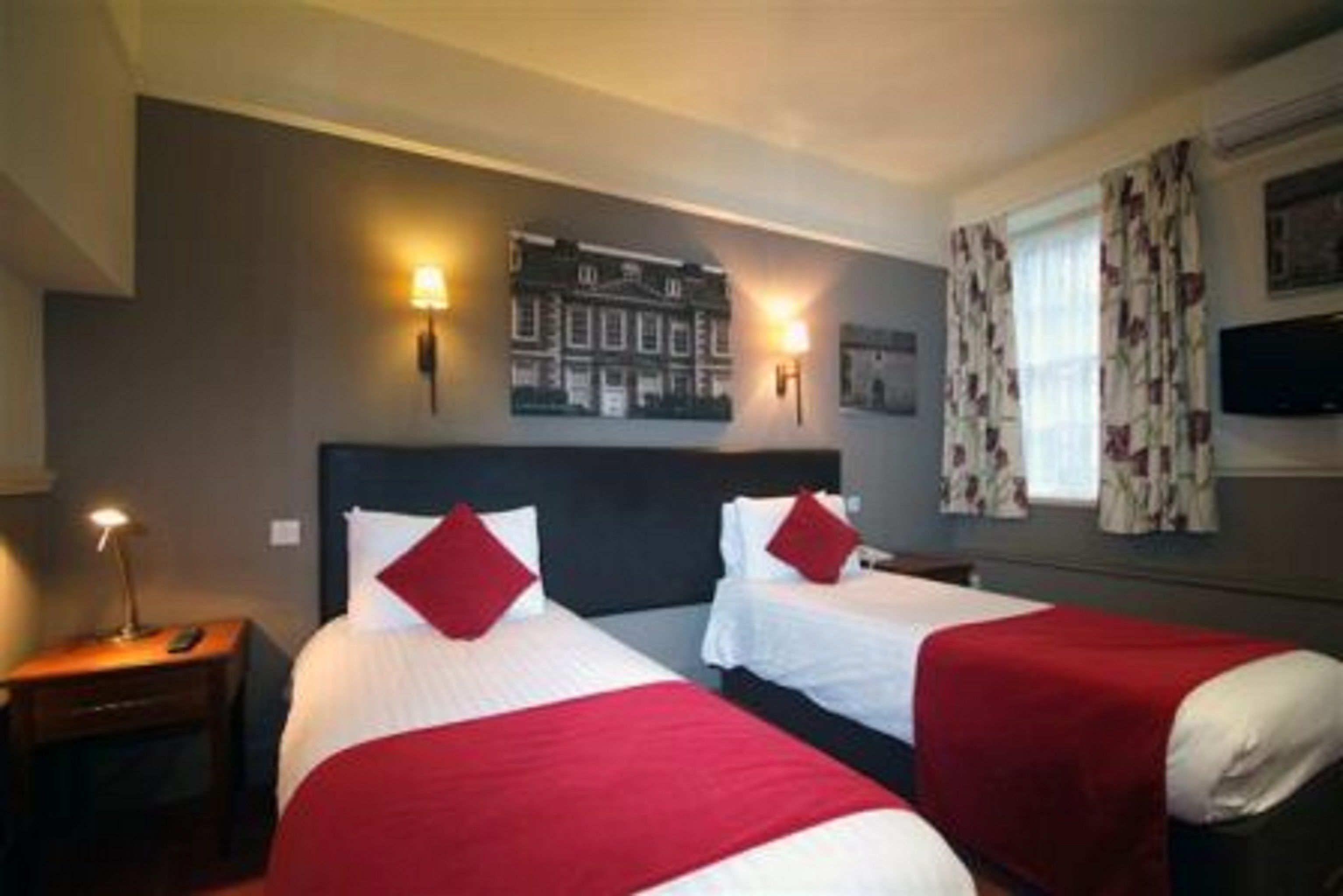 A hotel room with two beds, each with red and white bedding, a nightstand with a lamp, and a wall-mounted picture.