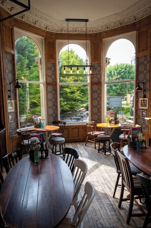 A cozy dining area with wooden tables and chairs, large arched windows with a view of greenery, and decorative wall patterns. - The Channings Hotel by Greene King Inns (Bristol)