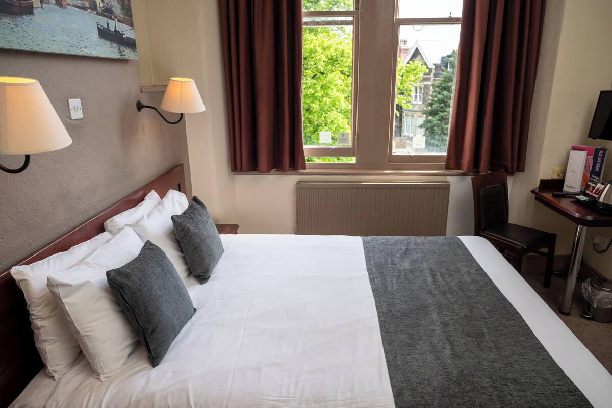 Een hotelkamer met een groot bed, twee wandlampen, een bureau met stoel, en een raam met gordijnen.