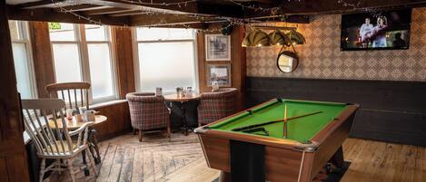 A pool table in a room with wooden floors, a billiards table, and a television mounted on the wall.