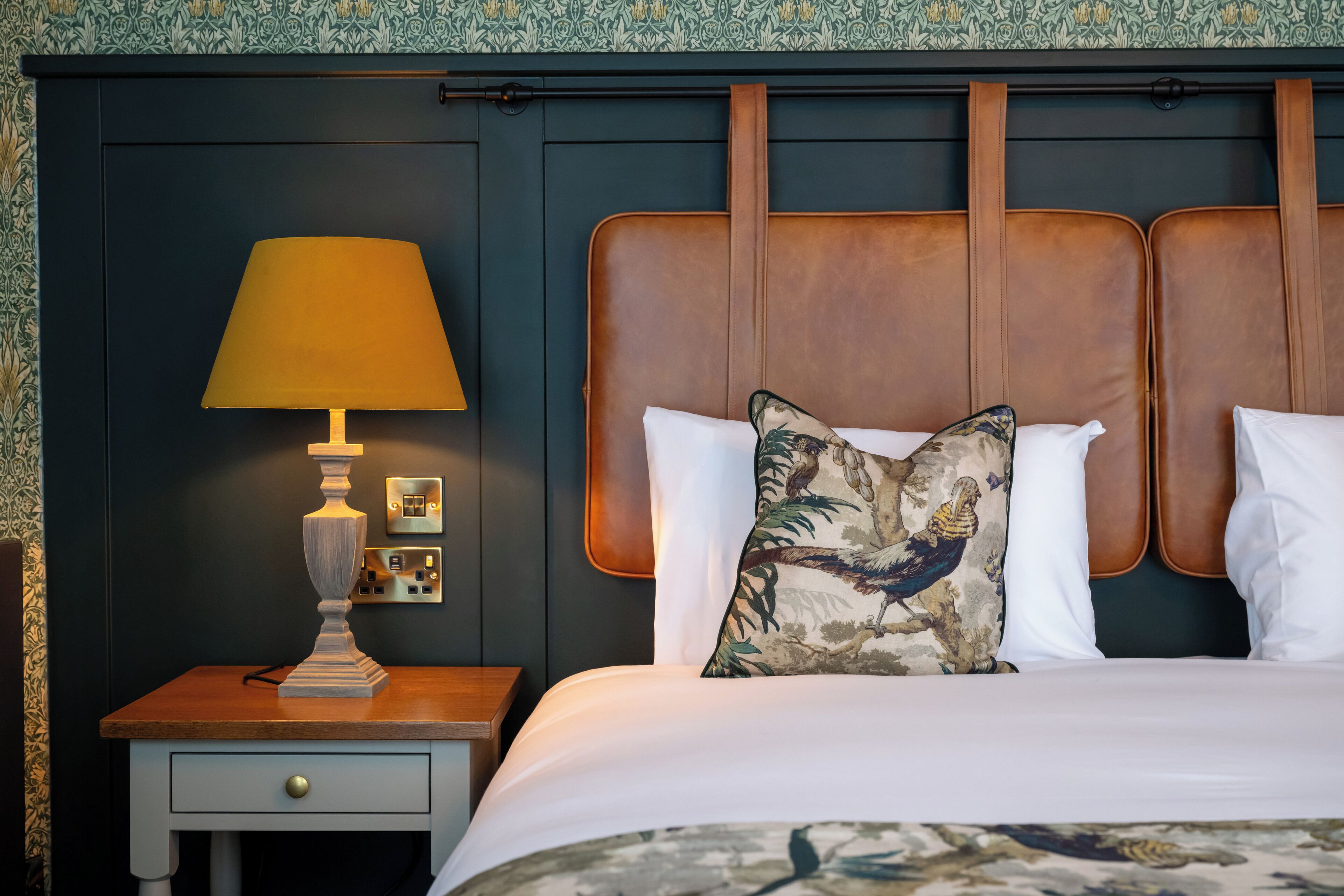 A neatly made bed with a headboard, a bedside table with a lamp, and a wall with a patterned wallpaper.