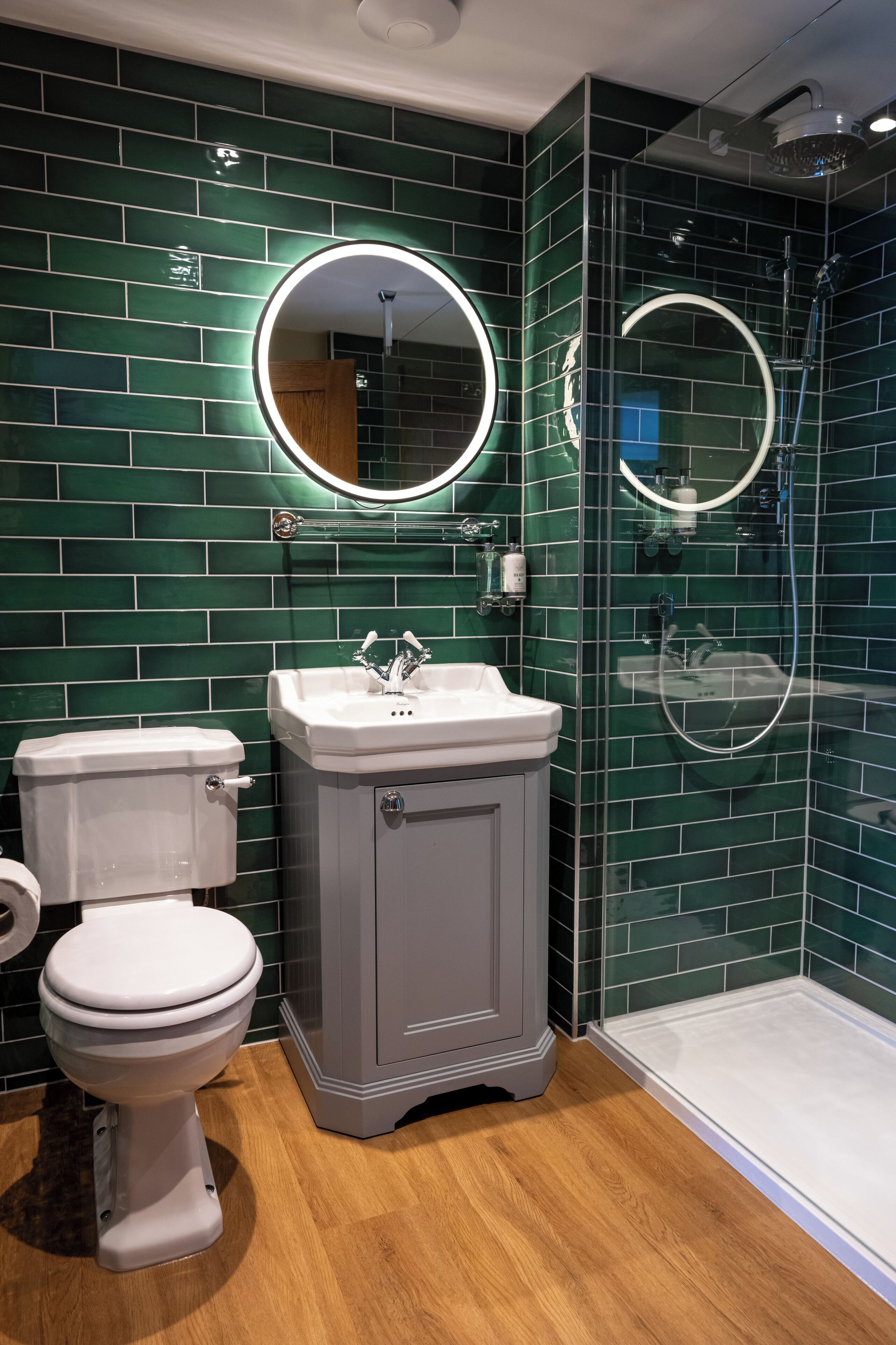 A bathroom with green-tiled walls, a white toilet, a sink with a mirror, and a glass-enclosed shower.
