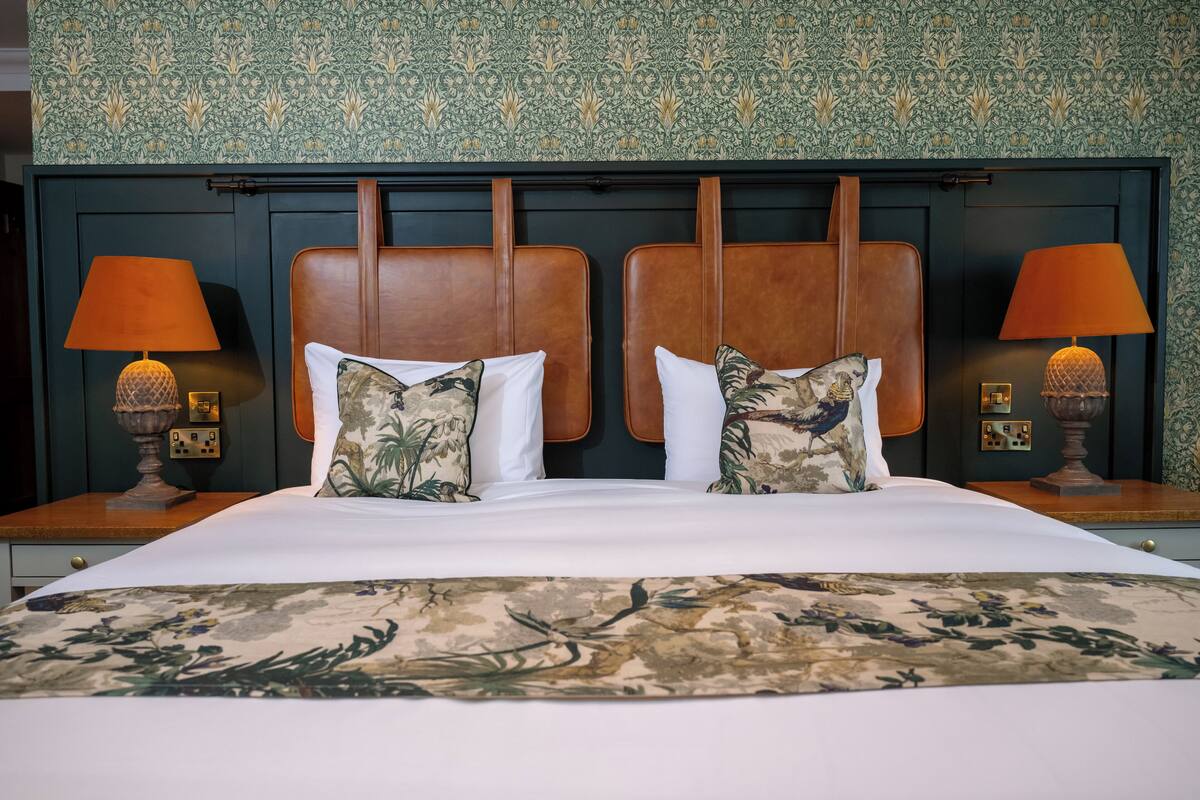 Un lit double avec une couvre-lit à motifs floraux, deux tables de chevet avec des lampes et une tête de lit ornée d’un papier peint à motifs.