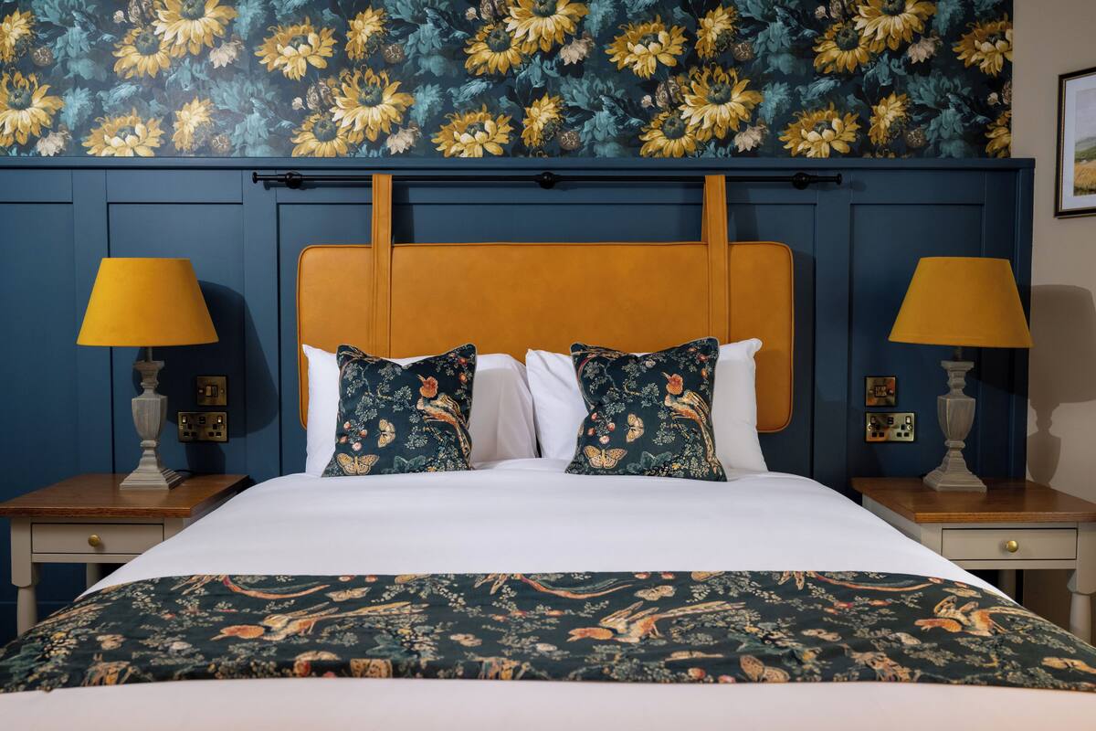 Un lit bien fait, agrémenté d’oreillers à motifs floraux et d’une couvre-lit assortie, flanqué de deux tables de chevet avec des lampes, le tout adossé à un mur recouvert d’un papier peint floral.