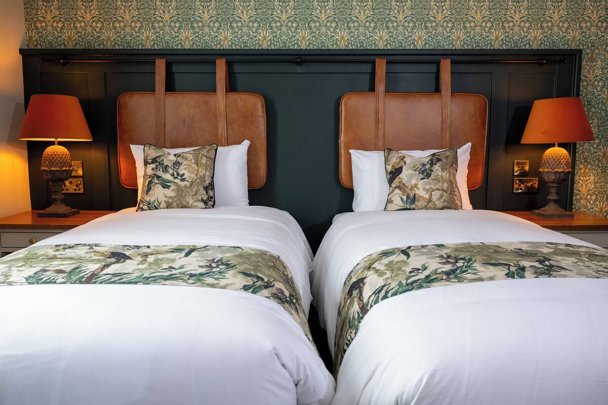 Une chambre d’hôtel avec deux lits, chacun recouvert d’une couvre-lit à motifs floraux et agrémenté d’oreillers assortis. Deux lampes de chevet, chacune munie d’un abat-jour, sont placées de chaque côté des lits.
