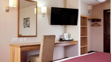 A hotel room with a wooden desk, a chair, a television, a bed, a mirror, and a shelf with a hanging coat.