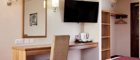 Habitación de hotel con un escritorio de madera, una silla, televisor, cama, espejo y estante con un abrigado colgado.