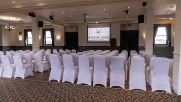 A conference room with rows of white chairs, a projector screen displaying "GREENE KING BURY ST EDMUNDS", and a chandelier.