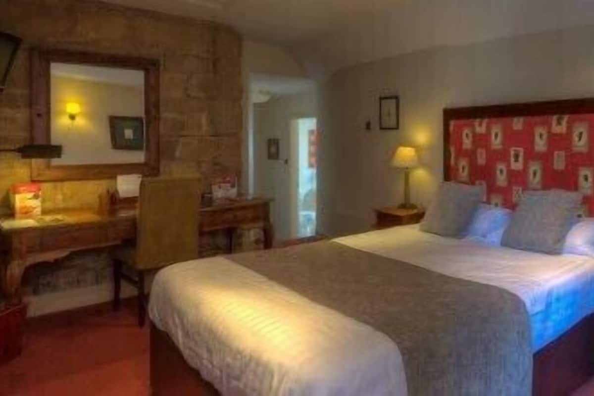 A hotel room with a large bed, a desk with a chair, a red armchair, and a wooden headboard.