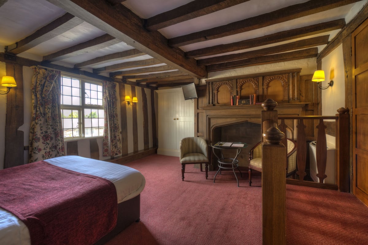 A traditional room with a bed, a chair, a small table, a fireplace, and a wooden staircase.