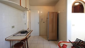 A compact kitchen with white tiled walls, a stainless steel refrigerator, a dining table with two stools, and a small window with an arched top. - Ribeiro 704 (Rio de Janeiro)