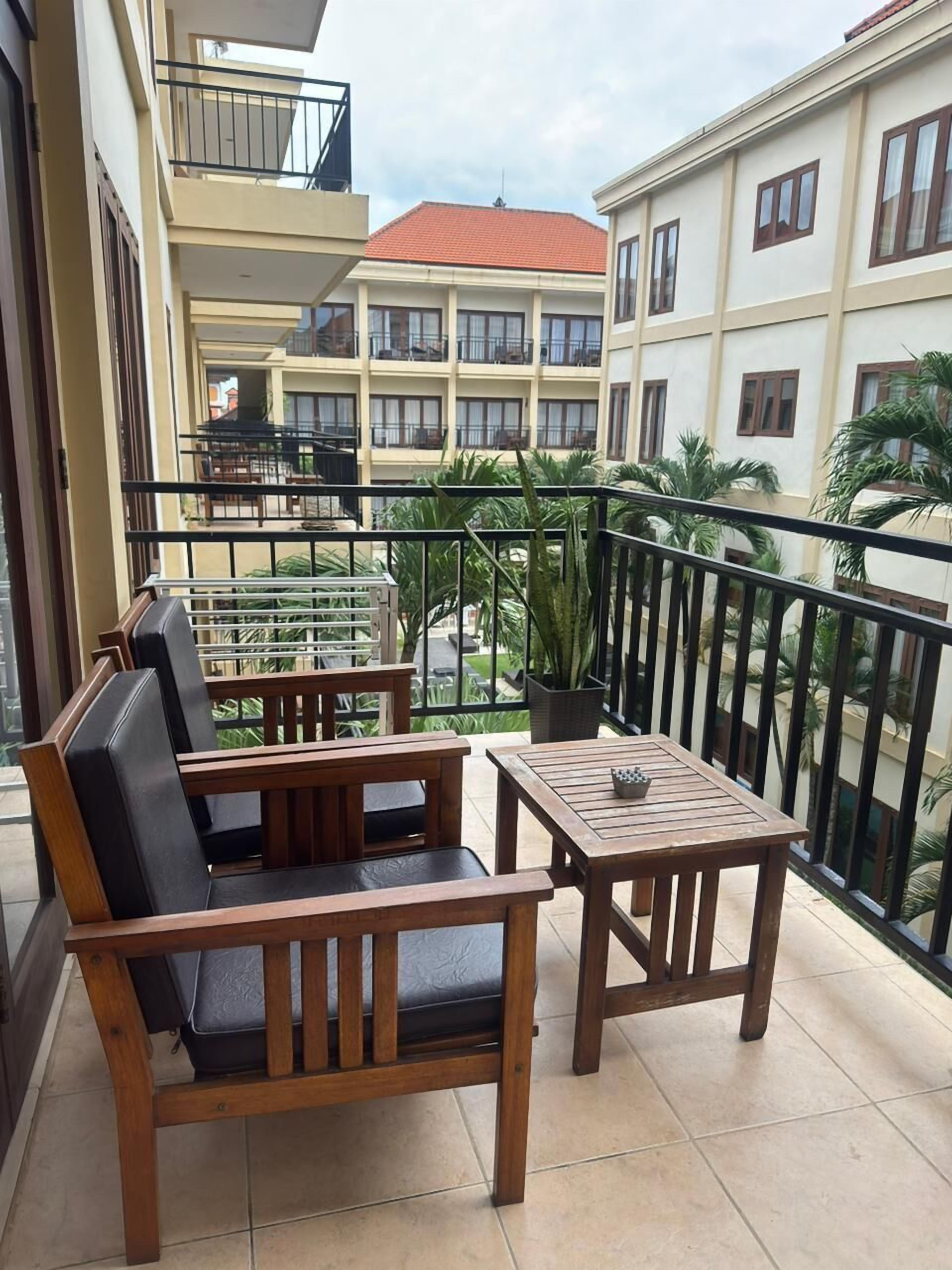 Un balcon doté de mobilier en bois, comprenant un canapé et une table, et offrant une vue sur un immeuble avec de nombreuses fenêtres et des balcons.