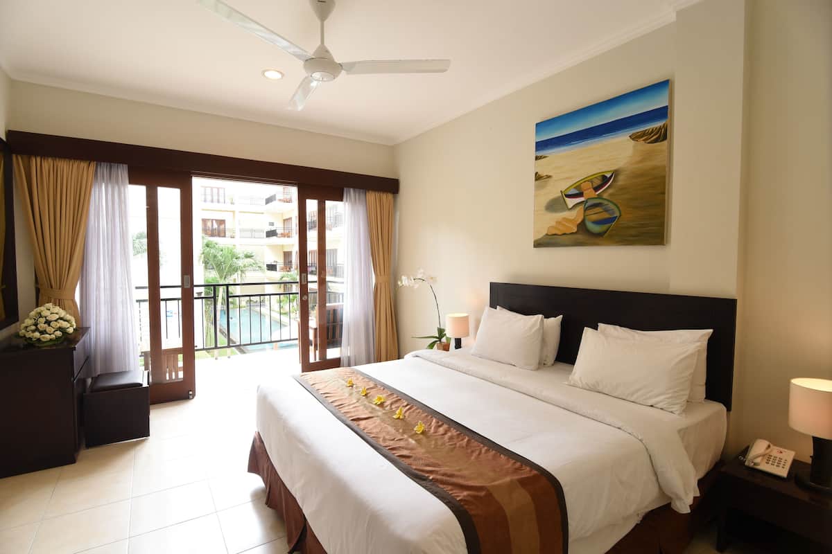 A hotel room with a large bed, a ceiling fan, a painting of boats on the beach, and a view of the outside balcony.