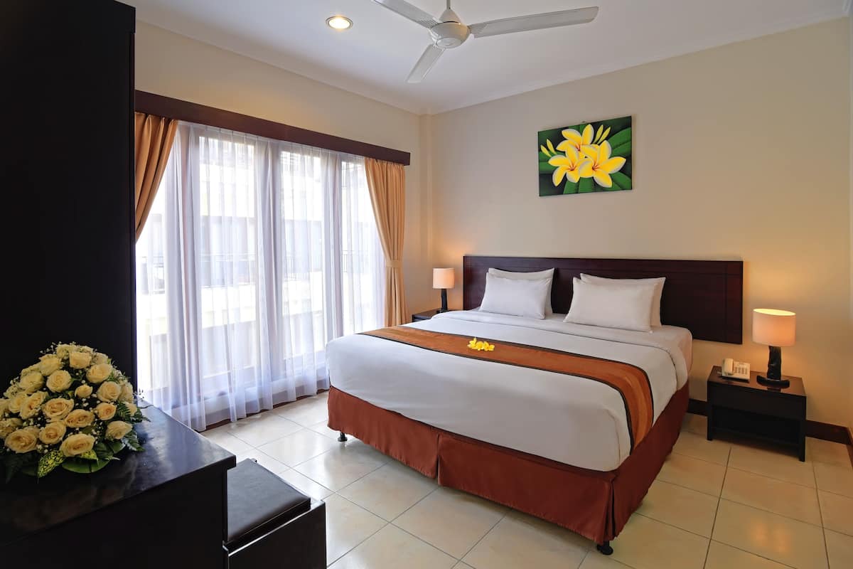 A hotel room with a large bed, a desk with a flower arrangement, a ceiling fan, and a painting on the wall.