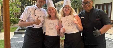 Een groep mensen in koksoutfits poseert met certificaten in een openlucht setting, bij een eettafel met traditionele gerechten.