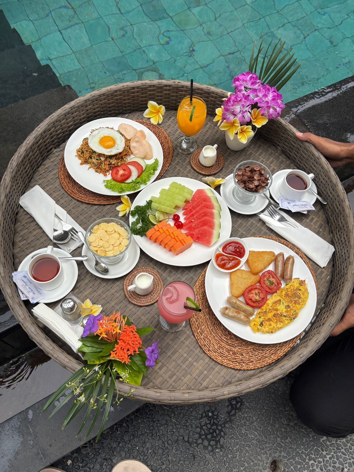 A tray with various breakfast items including eggs, toast, fruit, and beverages, placed on a woven mat near a pool.