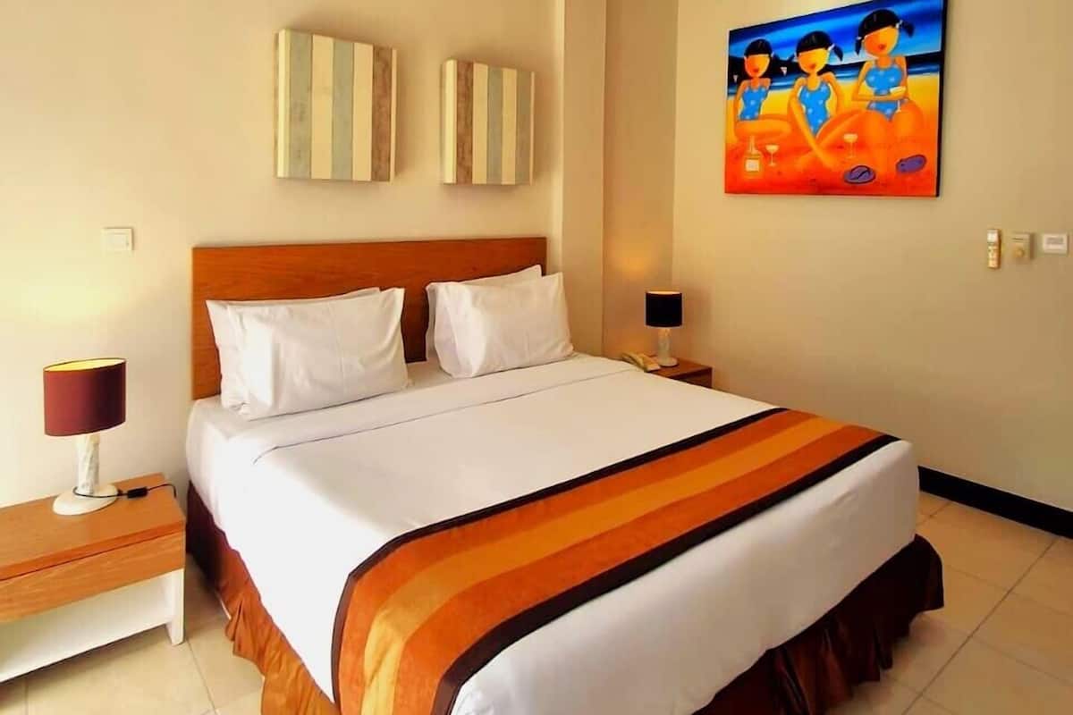 A hotel room with a bed, a ceiling fan, a painting, and a nightstand with a lamp.