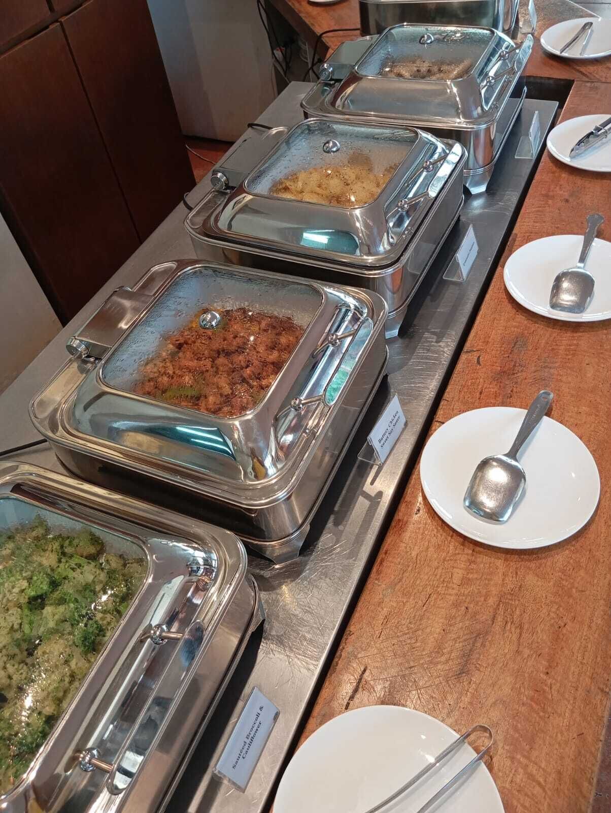 A buffet line with various food dishes in metal containers, accompanied by white plates and silver spoons.