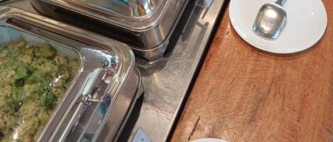 A buffet line with various food dishes in metal containers, accompanied by white plates and silver spoons.