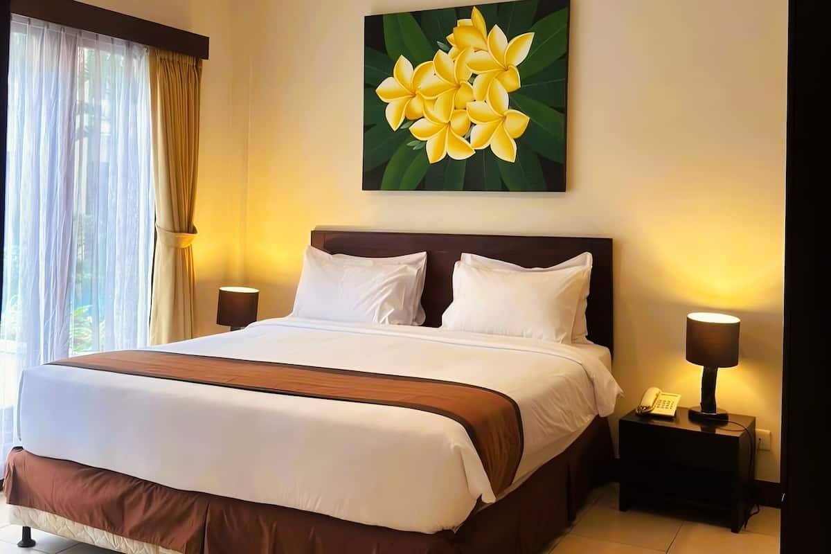 A hotel room with a bed, a ceiling fan, a nightstand with a lamp, and a painting of yellow flowers on the wall.