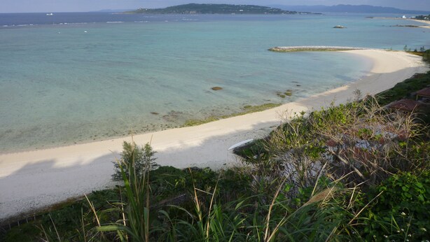 A coastal landscape with a sandy beach, clear turquoise waters, and a distant shoreline with vegetation and buildings. - Hibis Resort (Nakijin)