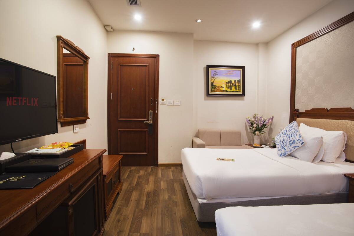 A hotel room with a wooden desk, a television, a bed with white bedding, a painting on the wall, and a door.