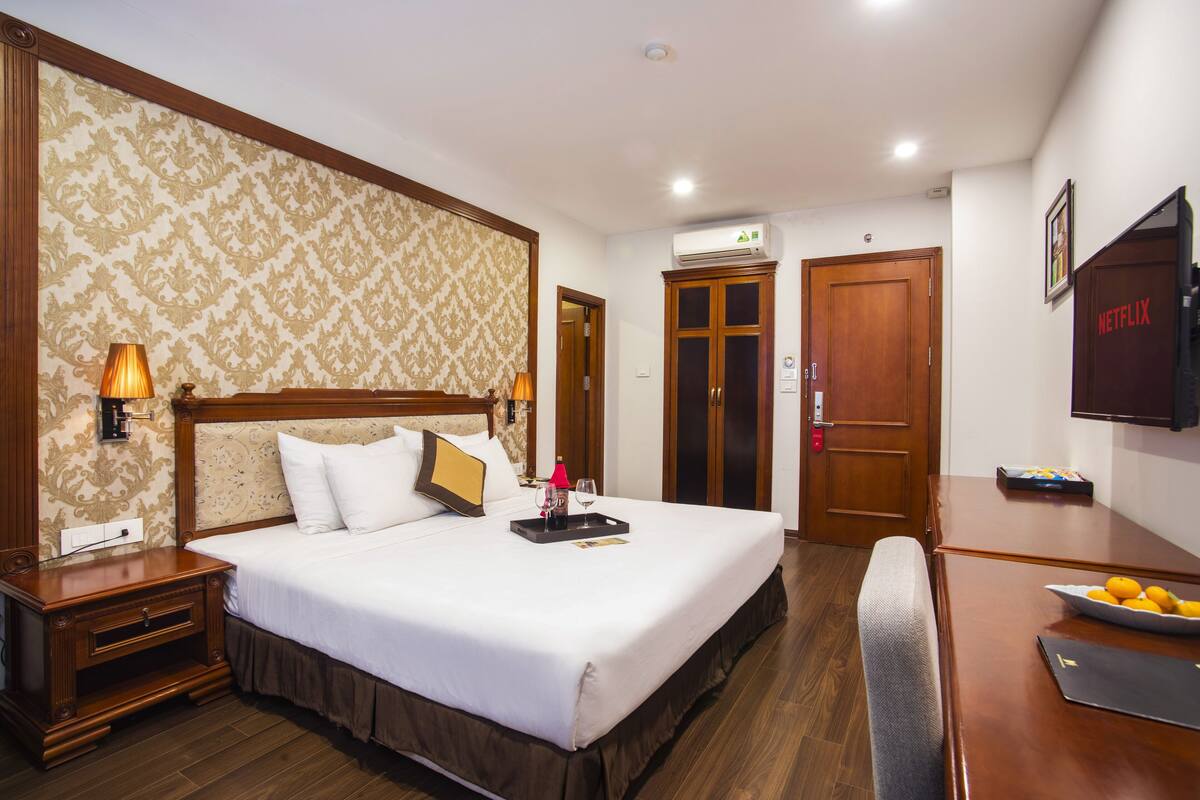 A hotel room with a large bed, wooden headboard, and a bedside table with a lamp. There is a desk with a laptop and a bowl of fruit.