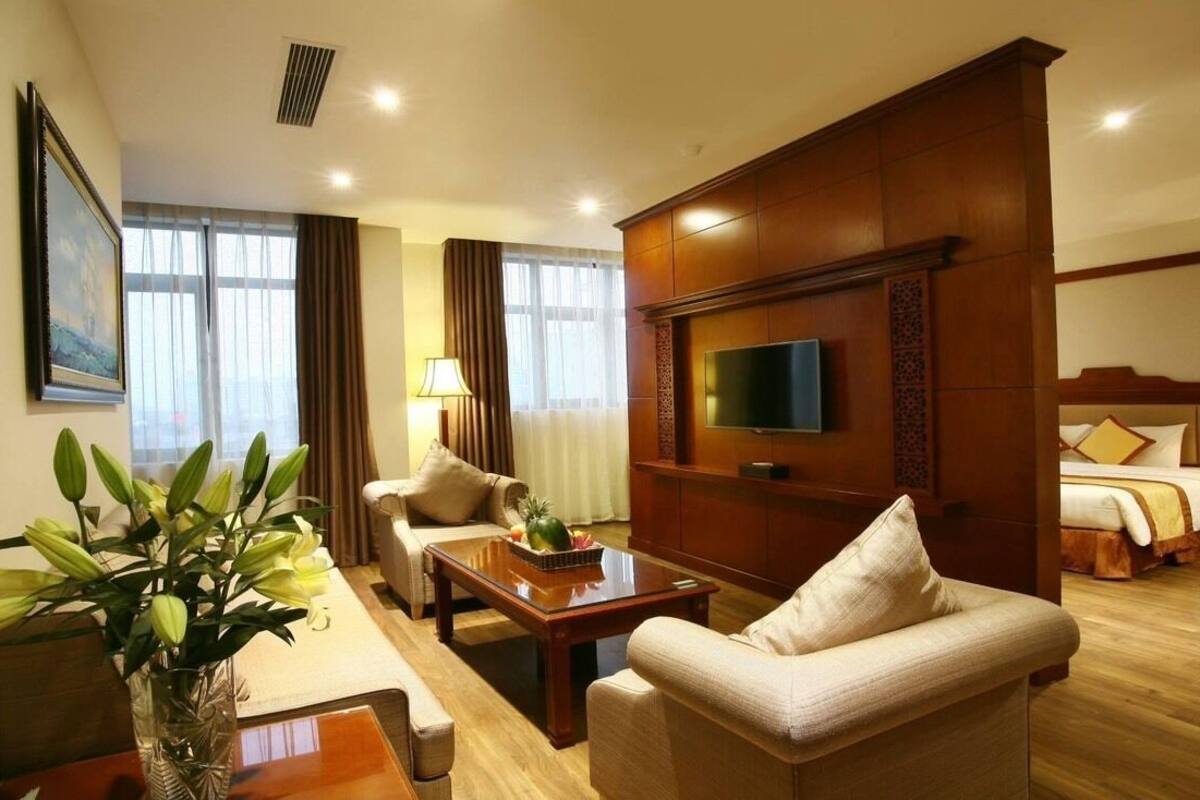 A hotel room with a large bed, a TV mounted on the wall, a wooden cabinet, a sofa, a coffee table, and a vase with flowers.