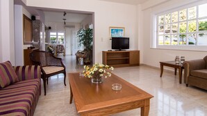 A living room with a sofa, a coffee table with flowers, a TV on a stand, and a window with a view of greenery. - Manolis Apartments (Hersonissos)