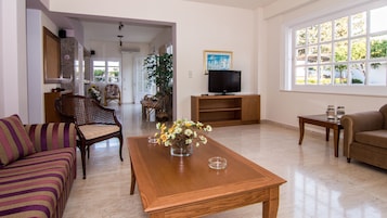 Un salón con sofá, una mesa baja con flores, televisor sobre un mueble y ventana con vistas a zonas verdes.