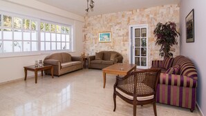 A spacious living room with a stone wall, a wooden coffee table, two sofas, a striped armchair, and a door leading outside. - Manolis Apartments (Hersonissos)