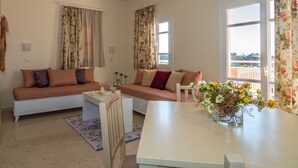 A cozy living room with a sofa and a small table, a window with curtains, and a view of the outside. - Manolis Apartments (Hersonissos)