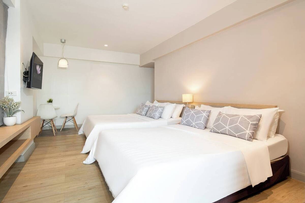 A hotel room with two beds, a desk, a chair, a TV, and a wall-mounted lamp.