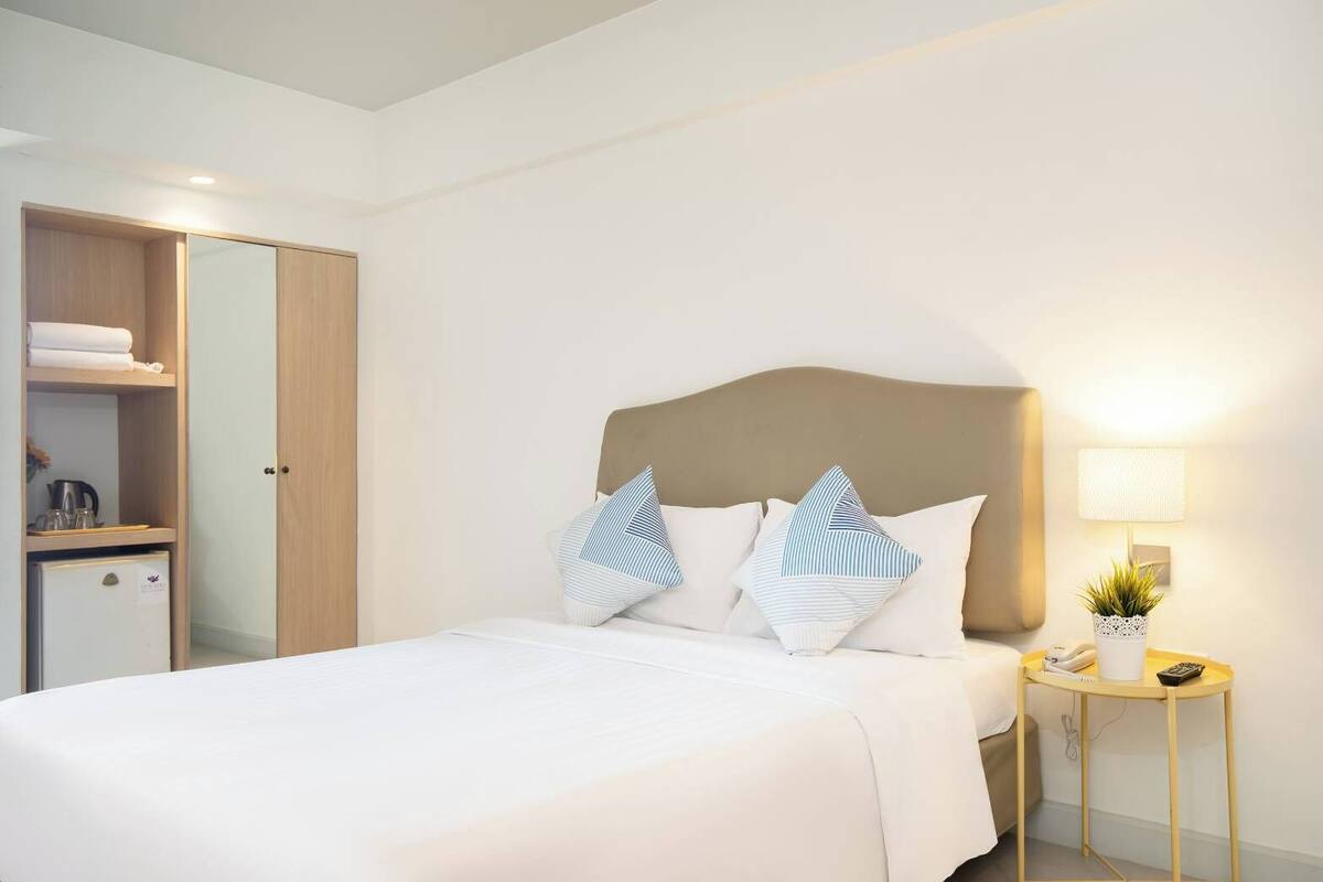 A neatly made bed with white bedding and striped pillows, a bedside table with a lamp and a small potted plant, and a wooden wardrobe with a mirror.