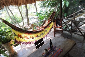 A hammock under a thatched roof, a wooden bench, a table with a bottle and glass, and a potted plant.