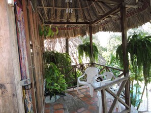 A covered outdoor seating area with white plastic chairs, a thatched roof, and surrounding greenery.