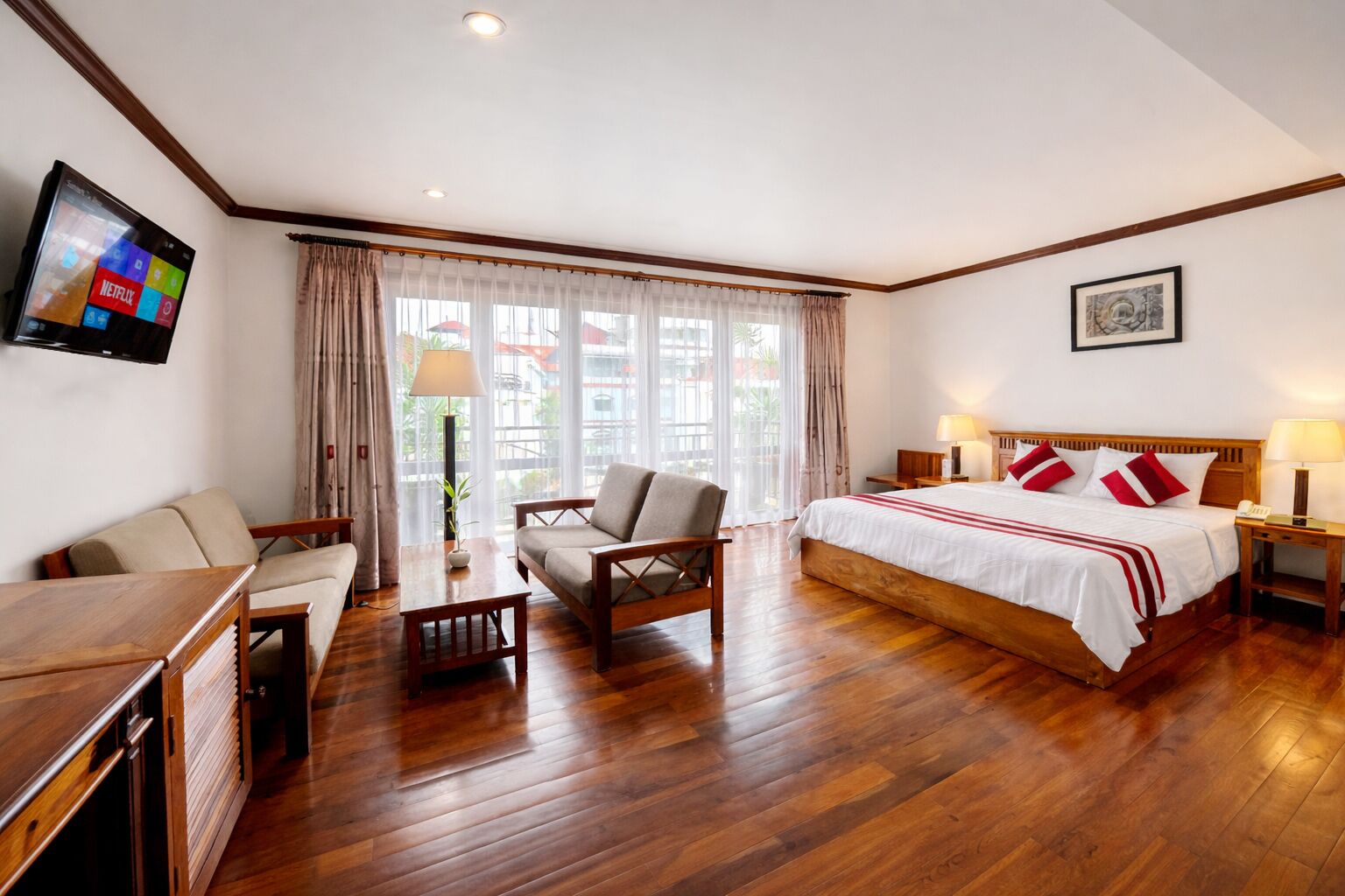 A hotel room with a wooden floor, a bed with red pillows, a TV mounted on the wall, and a window with curtains.
